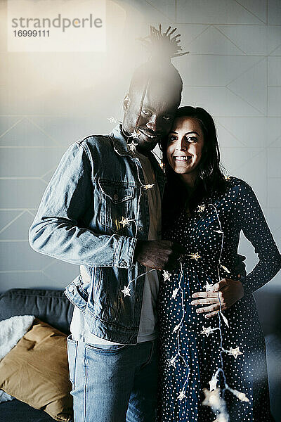 Glückliches multiethnisches Paar mit sternförmiger Beleuchtungsausrüstung  das zu Hause an der Wand steht
