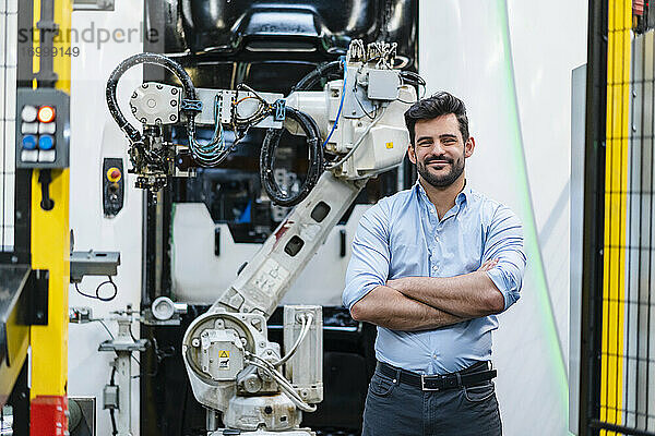 Lächelnder männlicher Unternehmer mit verschränkten Armen  der vor einem Roboterarm in der Industrie steht