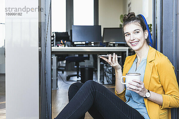 Geschäftsfrau mit Kaffeetasse  die mit ihrem Handy telefoniert  während sie am Eingang eines Büros sitzt