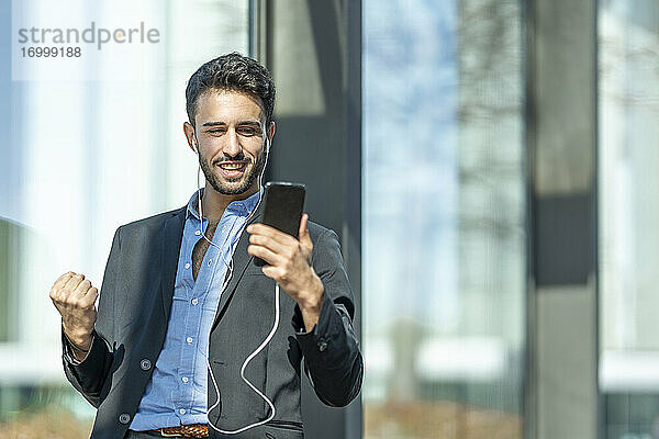 Lächelnder Geschäftsmann mit In-Ear-Kopfhörern  der jubelt  während er im Freien steht und ein Mobiltelefon benutzt