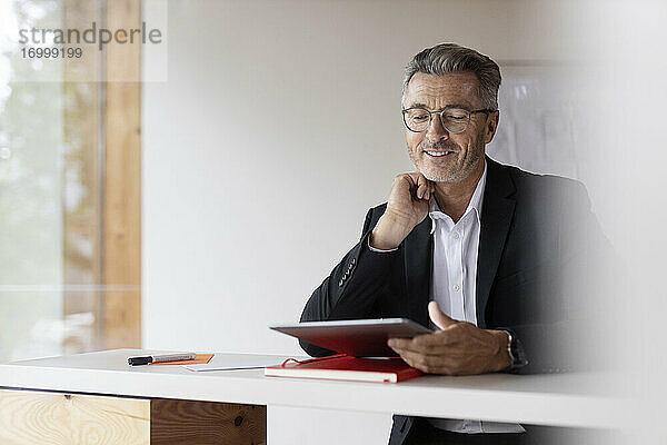 Lächelnder Geschäftsmann mit Hand am Kinn  der zu Hause ein digitales Tablet benutzt