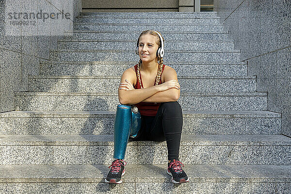 Lächelnde Sportlerin mit Beinprothese und Kopfhörern  die mit verschränkten Armen auf einer Treppe sitzt