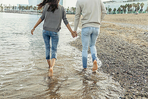 Junges Paar hält sich an den Händen  während es am Strand über das Wasser läuft