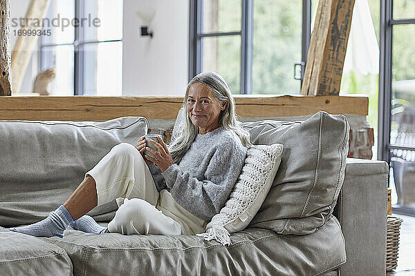 Lächelnde reife Frau hält eine Kaffeetasse  während sie zu Hause auf dem Sofa sitzt