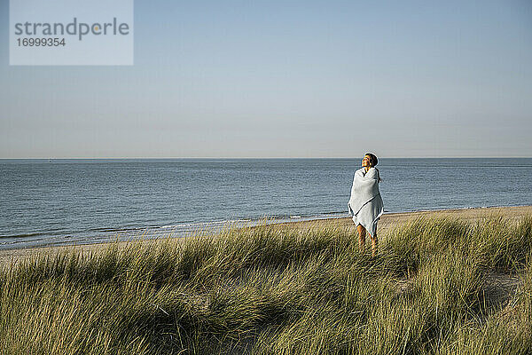 Junge Frau in Schal eingewickelt am Strand stehend