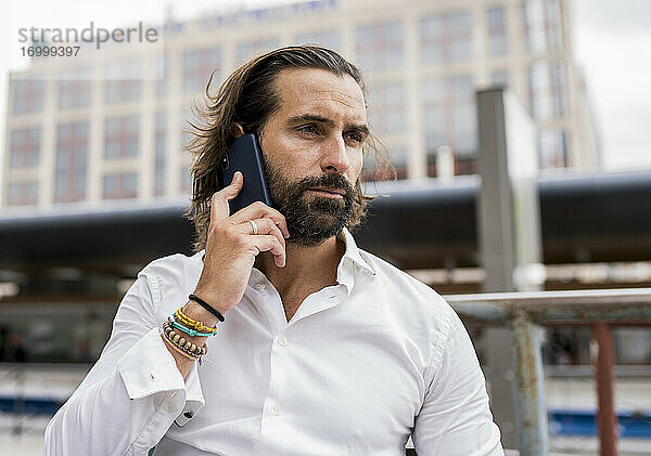 Unverfälschtes Porträt eines bärtigen Geschäftsmannes  der im Freien sitzt und mit seinem Smartphone telefoniert