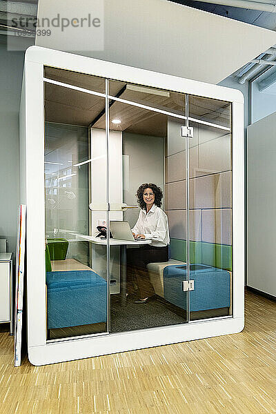 Lächelnde Geschäftsfrau  die einen Laptop benutzt  während sie in einer Kabine im Büro sitzt