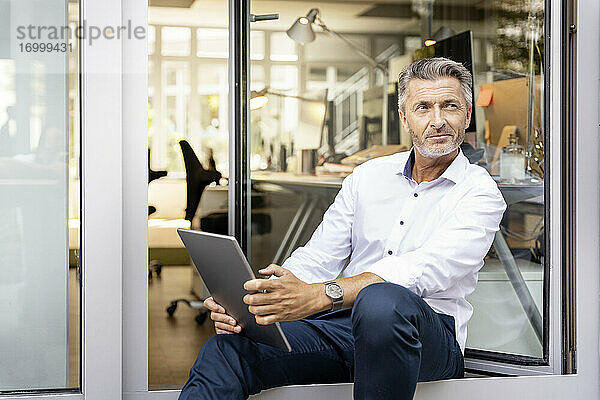 Nachdenklicher Geschäftsmann mit digitalem Tablet  der wegschaut  während er sich an die Bürotür lehnt