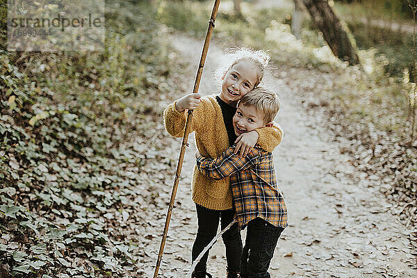 Schwester hält Stock und umarmt Bruder im Wald stehend