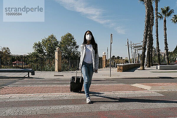 Frau mit Gepäck überquert Straße gegen blauen Himmel an einem sonnigen Tag während der Pandemie