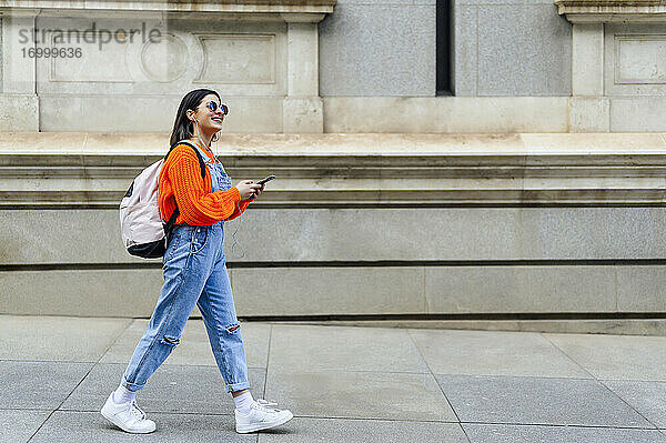 Lächelnde Frau mit Rucksack und Mobiltelefon  die beim Gehen auf dem Fußweg wegschaut