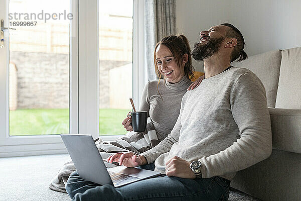Mann lacht  während er mit seiner Freundin zu Hause einen Film auf dem Laptop anschaut