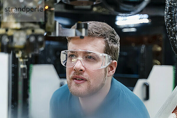 Männlicher Arbeiter mit Schutzbrille bei der Untersuchung eines Roboterarms in der Industrie