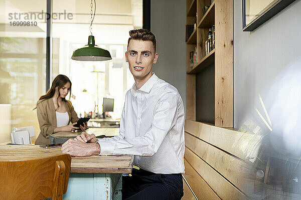 Junger Geschäftsmann sitzt auf einem Tisch mit einem Kollegen  der im Hintergrund im Büro arbeitet