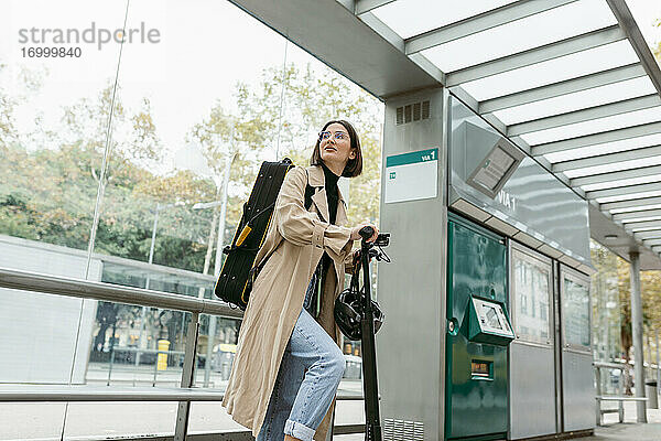 Lächelnde Frau mit Tretroller und Instrumentenkoffer an der Straßenbahn in der Stadt