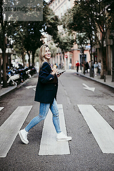 Lächelnde blonde Frau mit Handy in der Hand beim Überqueren der Straße in der Stadt