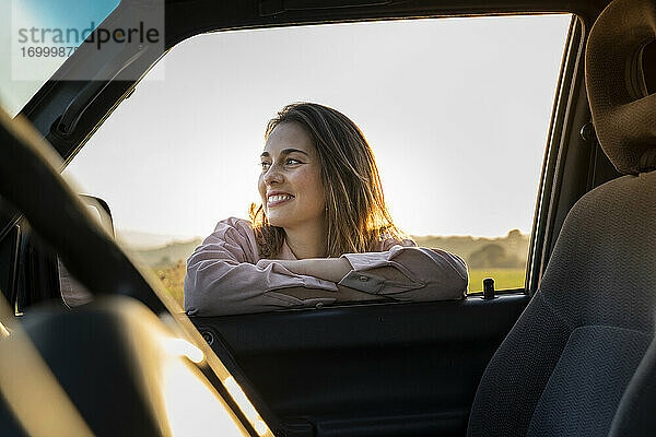 Glückliche junge Frau  die sich bei Sonnenuntergang an ein Autofenster lehnt und wegschaut