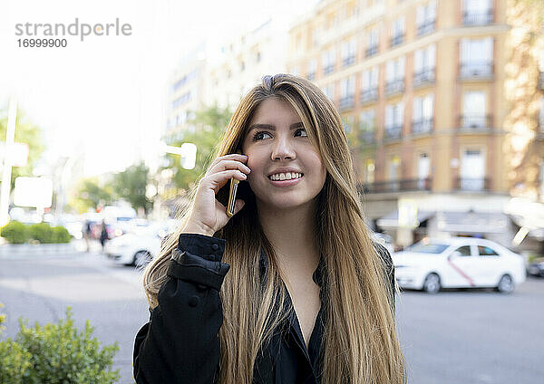 Lächelnde Frau  die auf der Straße in der Stadt steht und mit ihrem Handy telefoniert