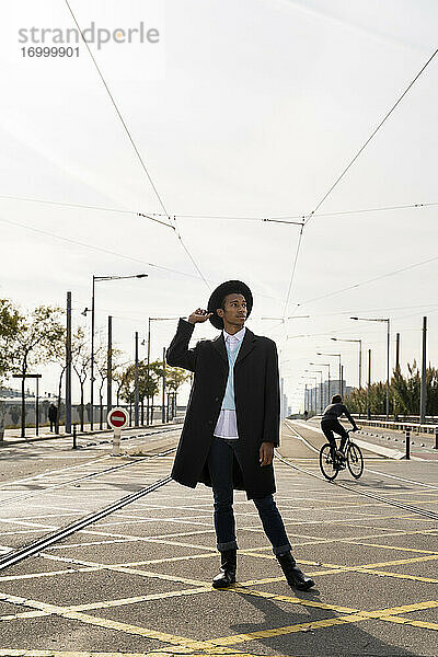 Junger Mann schaut weg  während er seinen Hut zwischen Eisenbahnschienen auf der Straße hält
