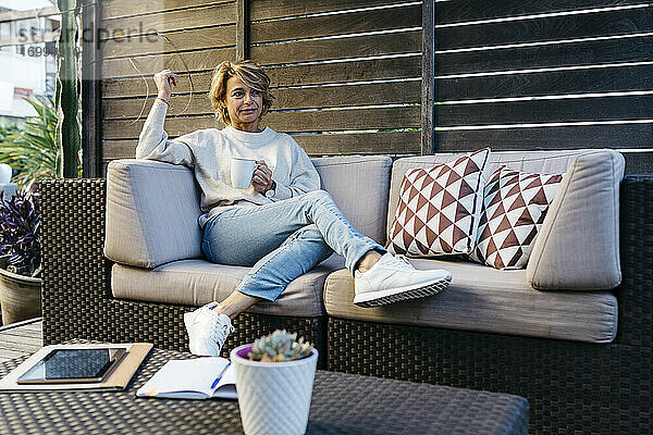Blonde Frau schaut weg und hält eine Kaffeetasse auf dem Sofa im Dachgarten