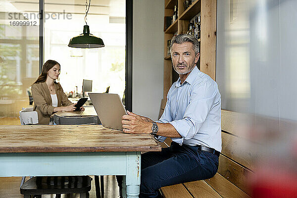 Geschäftsmann  der einen Laptop benutzt  während er mit einem Kollegen im Hintergrund im Büro sitzt