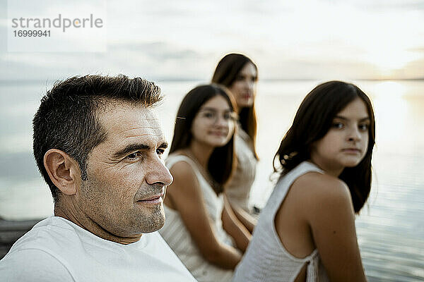 Vater sitzt mit Töchtern und Mutter am Steg am See