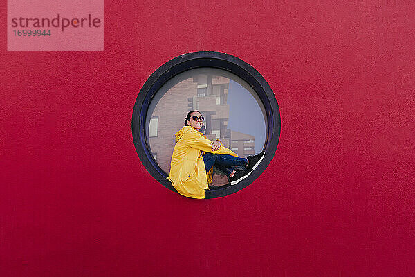 Frau in gelbem Regenmantel sitzt in Bullauge in roter Wand