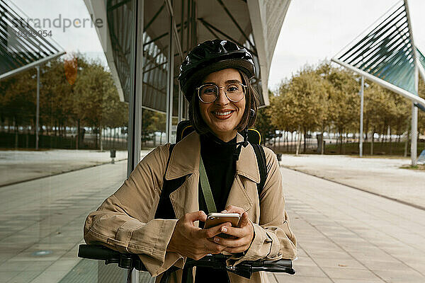 Lächelnde Frau mit Fahrradhelm  die mit einem Elektroroller auf dem Gehweg steht