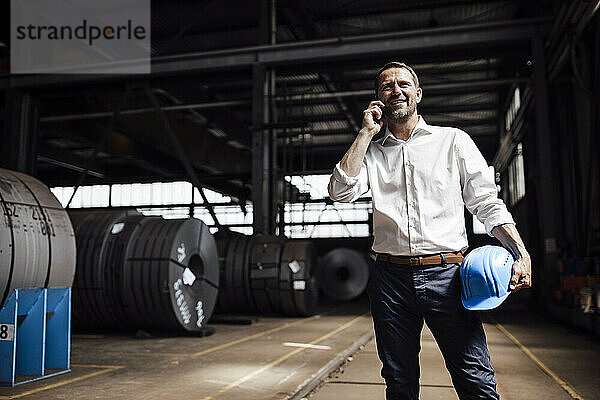 Lächelnder Geschäftsmann  der mit seinem Smartphone spricht  während er in der Industrie steht