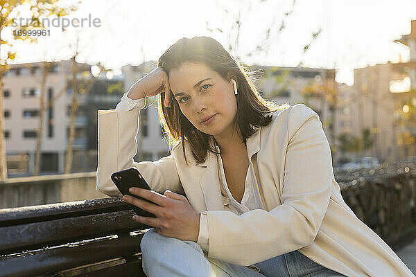 Nachdenkliche Unternehmerin hält ihr Smartphone in der Hand und sitzt auf einer Bank