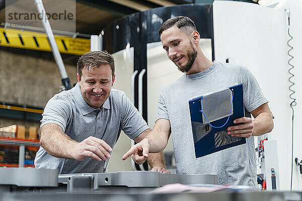 Älterer männlicher Techniker bei der Qualitätskontrolle eines Produkts  während er mit einem jungen Mitarbeiter in einer beleuchteten Fabrik steht
