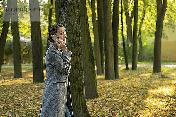 Weibliche Unternehmerin  die mit ihrem Smartphone telefoniert  während sie im Herbst im Park spazieren geht