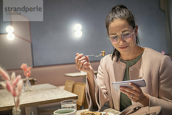 Lächelnde Geschäftsfrau  die etwas isst und dabei ihr Handy benutzt  in einem modernen Café sitzend