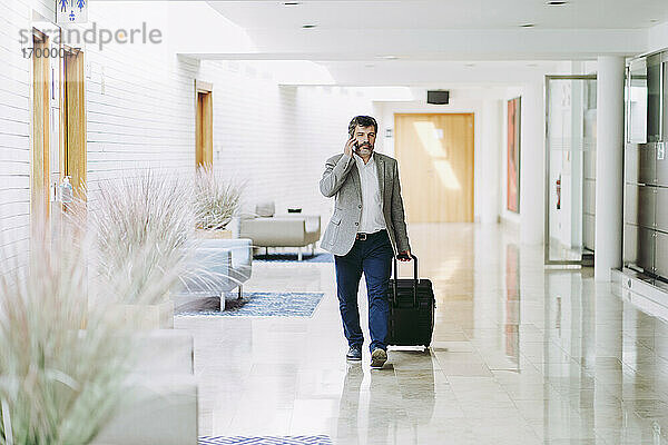 Männlicher Unternehmer am Telefon  der in einem Hotelkorridor ein Gepäckstück auf Rädern zieht