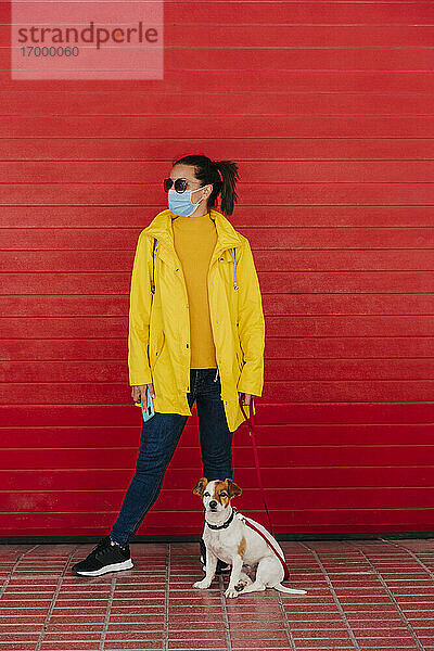 Frau mit Gesichtsmaske und Hund  in gelbem Regenmantel vor roter Wand