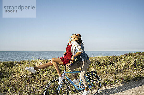 Glückliche Freundin sitzt auf dem Lenker  während sie eine Fahrradtour mit ihrem Freund gegen den klaren Himmel genießt