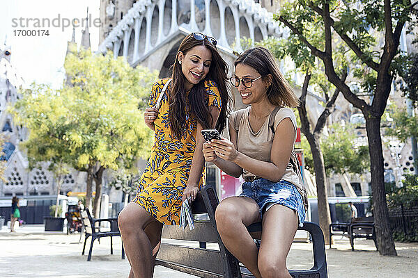 Frauen benutzen ein Mobiltelefon  während sie auf einer Bank vor der Sagrada Familia in Barcelona  Katalonien  Spanien sitzen