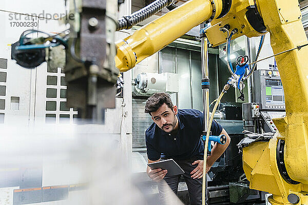 Männlicher Arbeiter mit digitalem Tablet  der einen Roboterarm in einer Fabrik untersucht