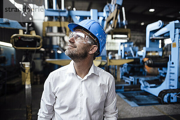 Lächelnder männlicher Unternehmer  der mit einer Schutzbrille in einer Fabrik nach oben schaut