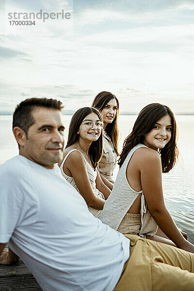 Glückliche Eltern sitzen mit ihren Kindern am Steg gegen See und Himmel