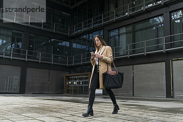 Modische Geschäftsfrau  die ein Mobiltelefon benutzt  während sie auf dem Fußweg vor einem Bürogebäude geht