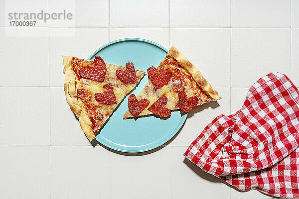 Pizza mit Peperoni in Herzform auf blauem Teller  weißen Fliesen und weiß-rot-karierter Serviette