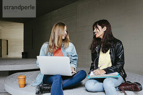 Lächelnde junge Freundinnen  die sich gegenseitig ansehen  während sie mit Büchern und Laptop auf dem Campus sitzen