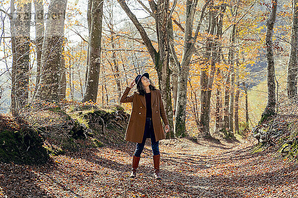 Frau mit Hut und Jacke  die auf einem Fußweg im Wald steht und nach oben schaut