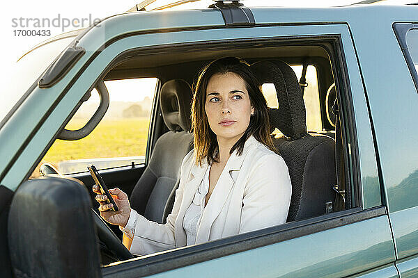 Junge Frau  die durch das Fenster schaut  während sie ihr Handy im Auto während einer Autofahrt hält