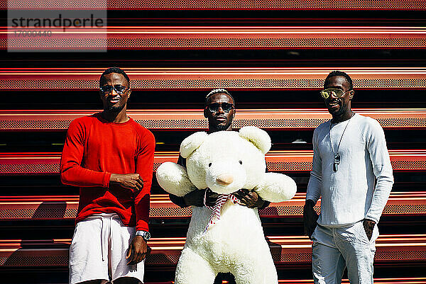 Porträt von drei coolen jungen Männern mit riesigem Teddybär an roter Wand