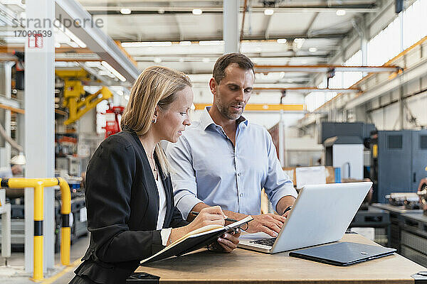 Unternehmerin mit Buch  während ein männlicher Kollege in einer Fabrik am Laptop arbeitet