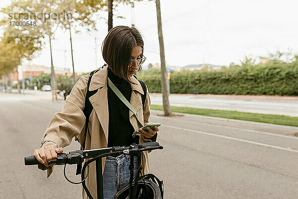 Mittlere erwachsene Frau  die ein Mobiltelefon benutzt  während sie mit einem Elektroroller auf der Straße steht