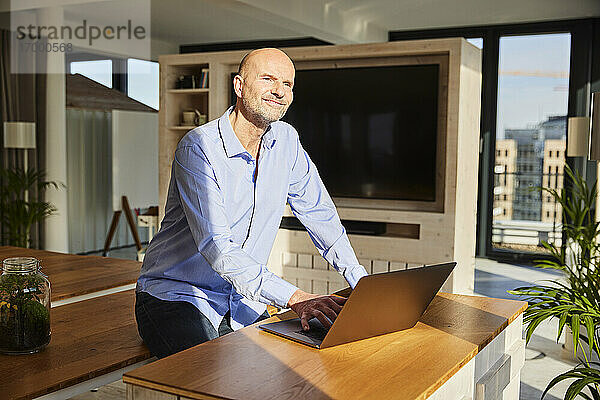 Lächelnder Geschäftsmann  der wegschaut  während er einen Laptop benutzt und zu Hause sitzt