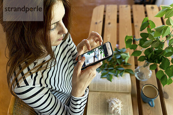 Junge Frau fotografiert Kaffeetasse und Buch auf dem Tisch mit dem Handy  während sie zu Hause sitzt